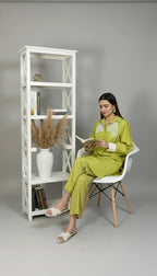 Woman in a green outfit reading a book next to a white bookshelf against a gray background