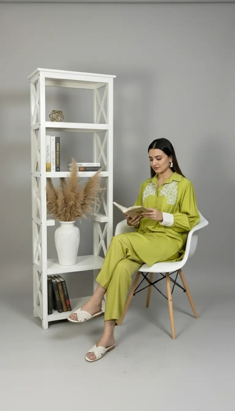 Woman in a green outfit reading a book next to a white bookshelf against a gray background