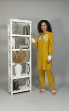 Woman in mustard yellow outfit standing next to a white bookshelf against a gray background